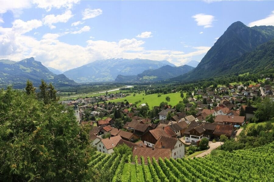 A green valley in Lichtenstein with a small town of orange-roofed houses and a backdrop of mountains.