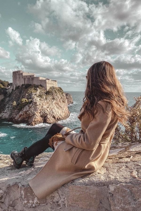 A woman sitting on the walls of Dubrovnik looking out at the Lovrijenac Fortress and Adriatic Sea.